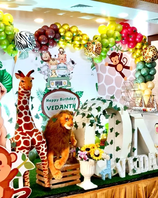 A close-up of a vibrant jungle safari theme for a first birthday. The setup includes a large "ONE" sign wrapped in vines, plush lions and giraffes, and a multi-colored organic balloon arch with zebra and leopard print accents.