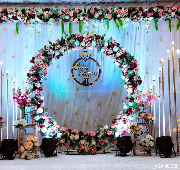 A stunning engagement stage centered around a large, lush floral ring made of pink and white roses. A custom sign with a ring icon hangs in the middle, all set against a beautifully lit white drape backdrop for a classic and grand celebration.