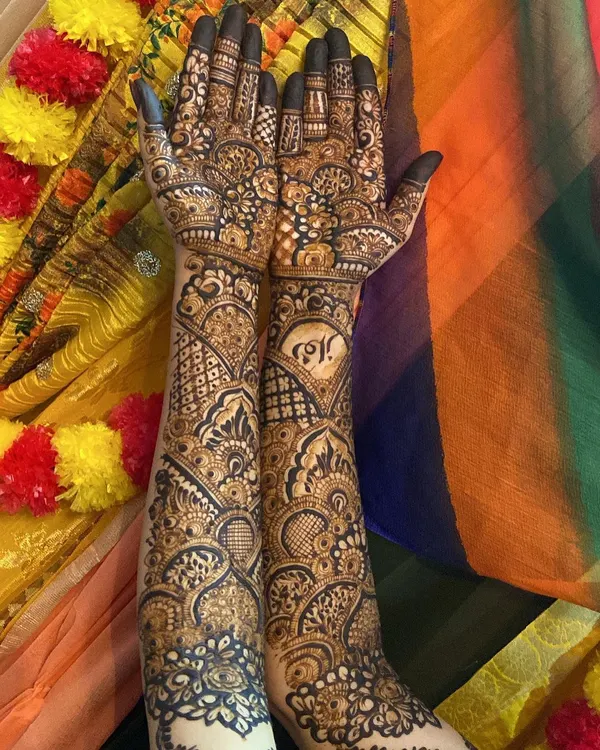 A complete bridal look featuring personalized initials within a heart on one arm. The design is dense with a mix of geometric grids, floral patterns, and traditional motifs, covering the arms fully up to the elbow.