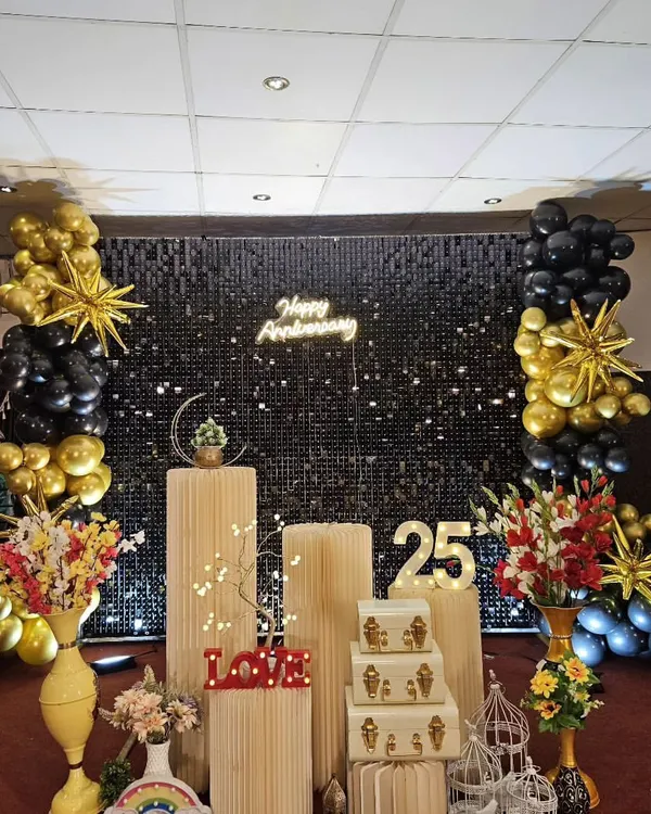 A sophisticated 25th anniversary decoration featuring a black shimmer wall and balloon columns in a chic black and gold color scheme. The setup includes a 'Happy Anniversary' neon sign and illuminated '25' marquee numbers.