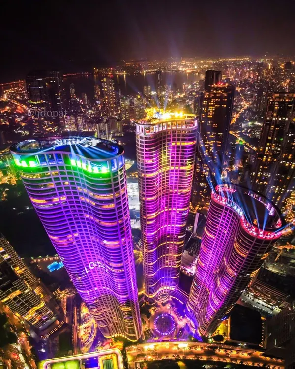 An aerial drone view captures the World One towers in Mumbai, illuminated with vibrant purple and green lights that paint the night skyline.