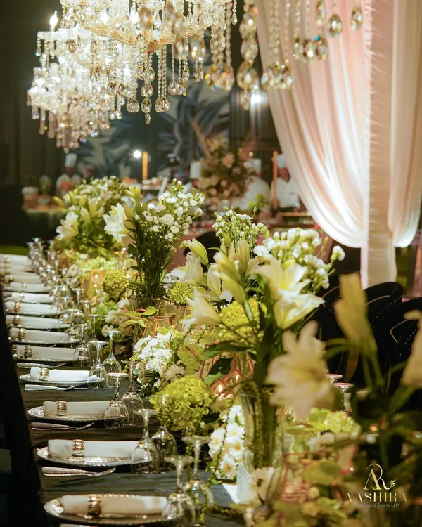 A long dining table set for a sangeet, with a runner of white lilies and green hydrangeas, illuminated by the glow of crystal chandeliers.