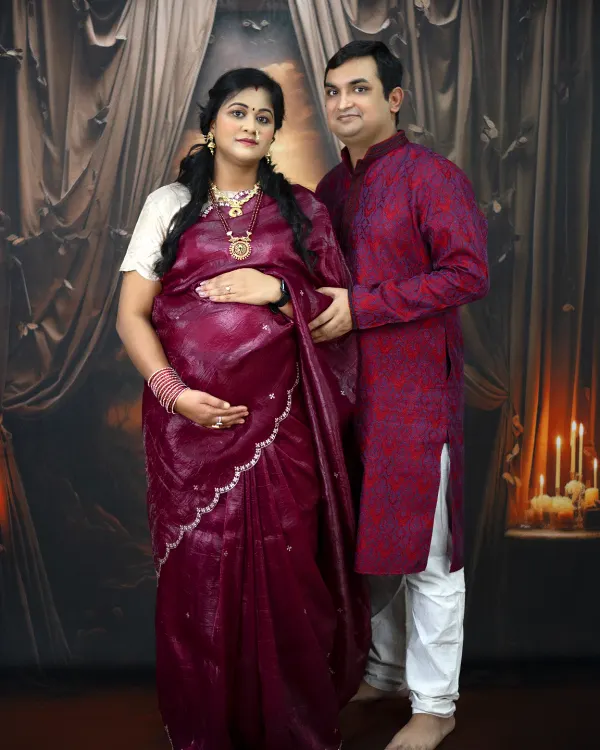 A glamorous and traditional maternity portrait. The couple is dressed in beautiful ethnic wear, posing against a classic, candlelit backdrop for a regal feel.