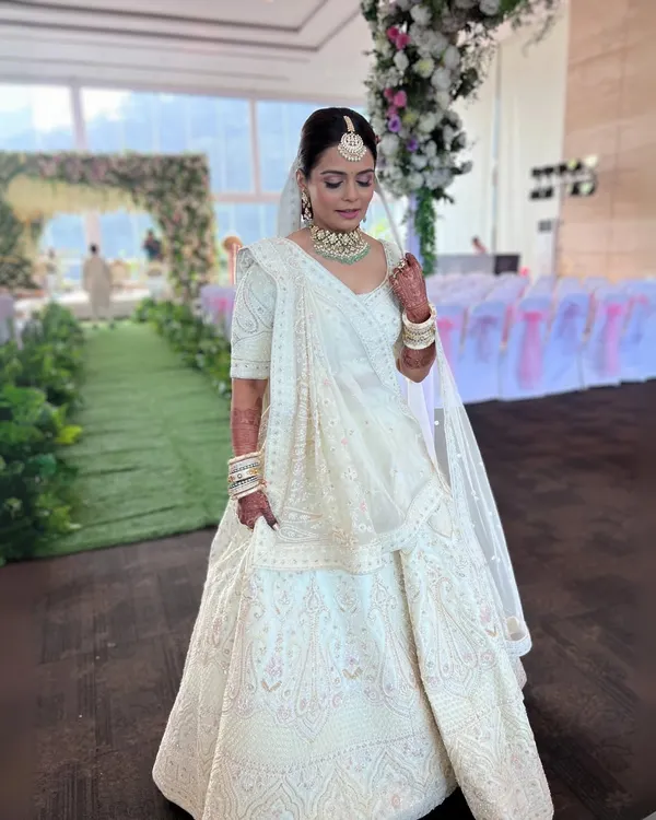 A breathtaking full-length shot of a bride in an all-white lehenga. The makeup is kept minimal and regal, with a clean bun adorned with velvet flowers, proving that elegance lies in simplicity.