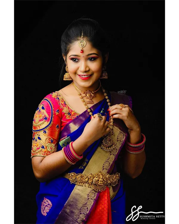 A classic South Indian bridal style from a model shoot, featuring a vibrant blue and pink Kanjeevaram saree. The makeup is bright and celebratory, with defined eyes and a cheerful lip color.