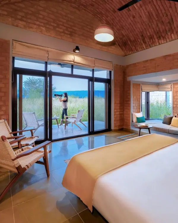 This is a bedroom in the Waghoba Ecolodge, a space designed for tranquility and immersion in nature. The vaulted ceiling, made from conical pottery tiles, and walls of stabilized adobe blocks create a cool, earthy sanctuary, while the large window opens directly to the restored forest landscape of Tadoba.