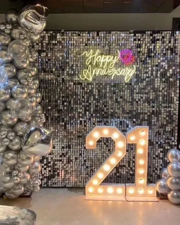 Celebrate your milestones in style. This chic 21st anniversary setup features a shimmering silver sequin wall, a custom "Happy Anniversary" neon sign, and large light-up marquee numbers for a glamorous photo corner.