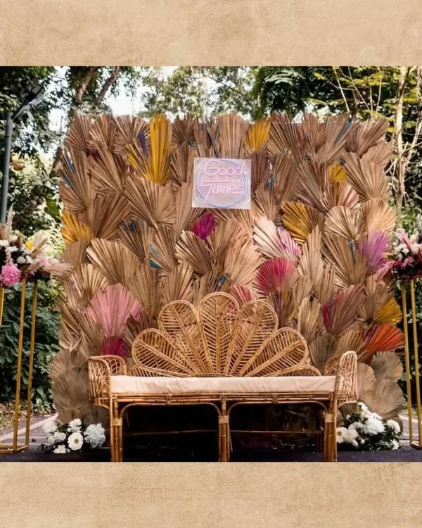 A cozy and stylish couple seating area with a boho theme. We used a beautiful peacock-style rattan bench set against a backdrop of dried and colored palm leaves, with a "Good Times" neon sign.