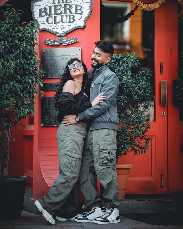 A fun and candid pre-wedding shot of a couple in casual, coordinated outfits in front of a vibrant red doorway at The Biere Club in Bangalore.