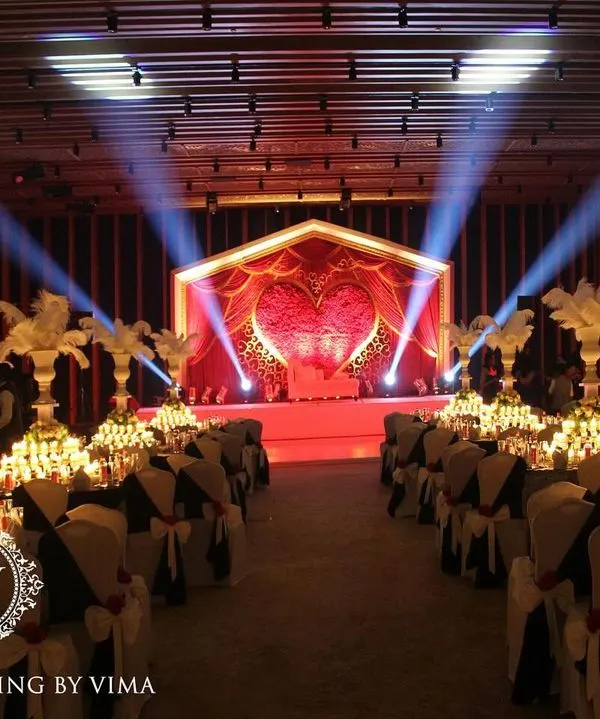 A glamorous anniversary celebration with a dramatic red and black theme. The room is filled with candlelit tables, large white feather centerpieces, and a stunning stage with a heart-shaped light installation.
