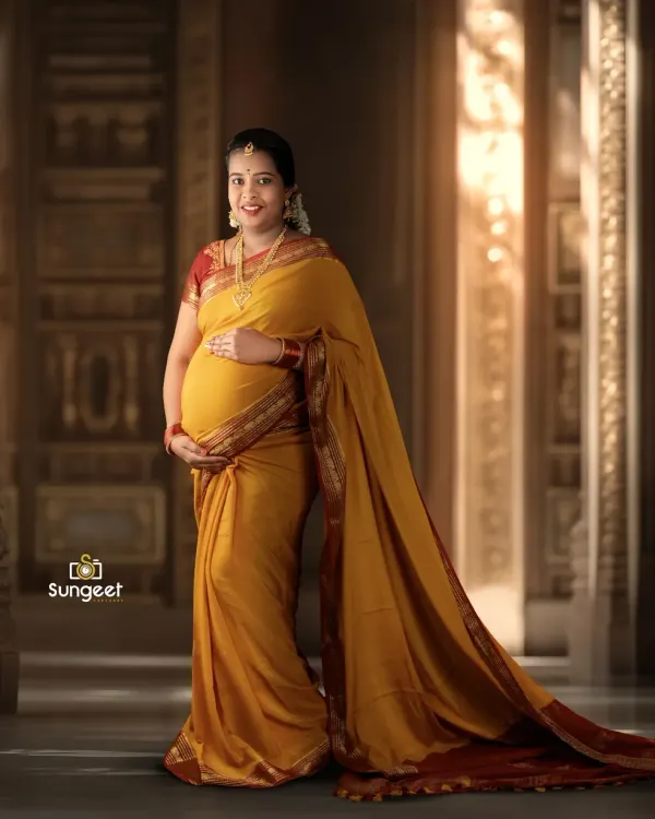 A full-length portrait showcasing the beautiful drape of a yellow and red silk saree. The architectural backdrop and soft lighting add a touch of grandeur to this traditional maternity photo.