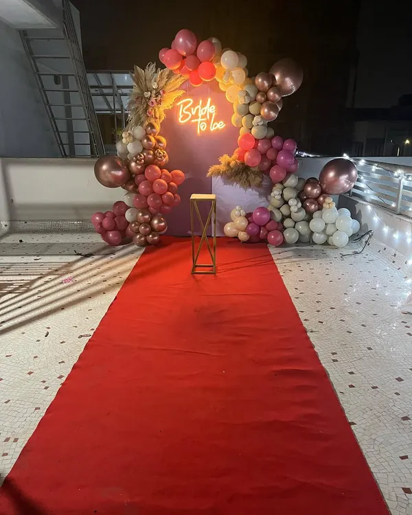 I create a grand entrance for the bride-to-be with a classic red carpet walkway. This setup features a custom backdrop with a 'Bride to Be' neon sign and a balloon arch in rose gold and pink tones, perfect for making her feel special.