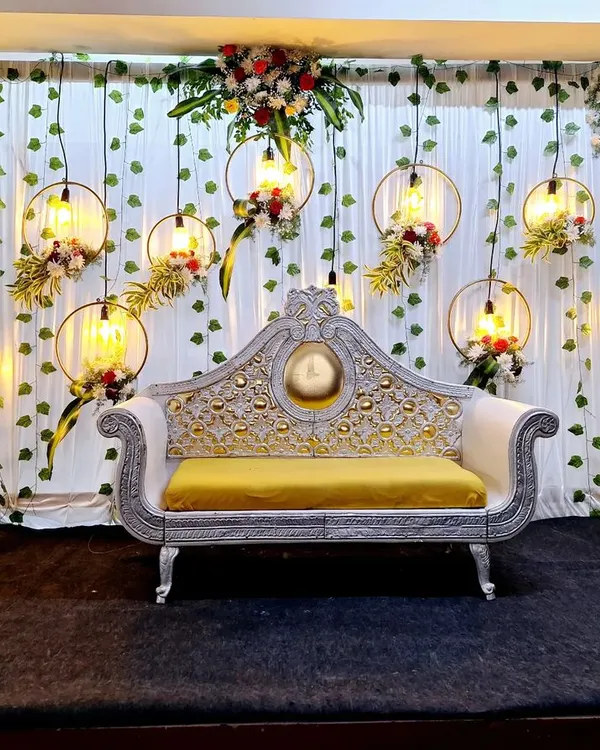 This reception stage combines rustic and industrial elements, featuring hanging floral hoops with exposed Edison bulbs. The design is set against a backdrop of green vines and white drapes, creating a warm, romantic glow.