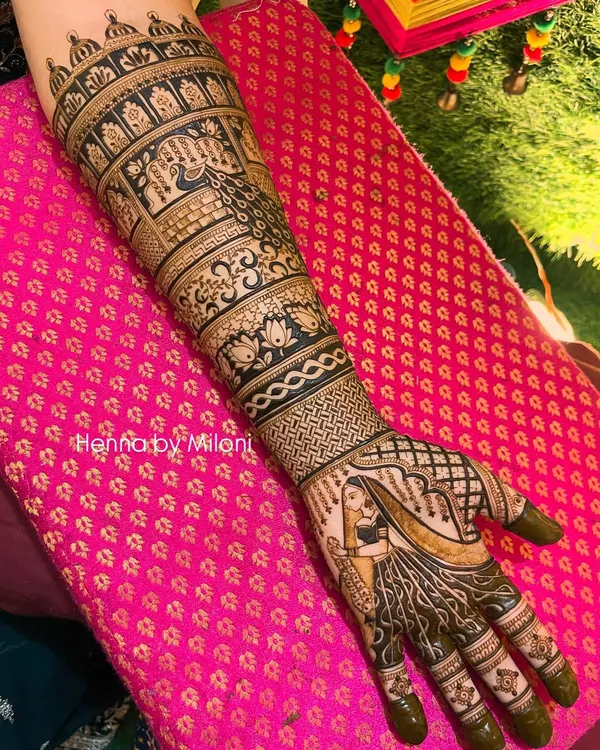 A full-arm bridal mehendi design showcasing a beautiful bride figure. The entire arm is covered in layers of traditional patterns, including peacocks, lotuses, and intricate jaal work, creating a truly magnificent look.