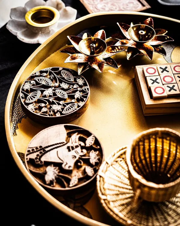 A curated tray of artisanal gifts, showcasing the beauty of traditional craftsmanship. It features hand-carved wooden printing blocks, brass lotus candle holders, and a wooden tic-tac-toe game, perfect for a memorable guest welcome hamper.