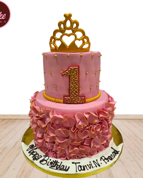 A stunning two-tiered princess cake for a first birthday. This pink and gold creation features delicate ruffles, a large number '1', and a golden crown topper for a royal celebration.