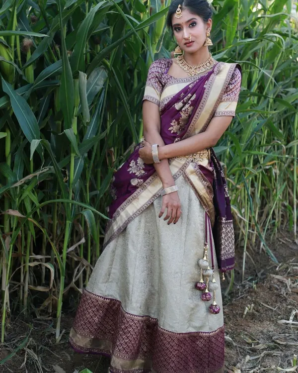 A full-length photo of the purple and cream Langa Voni set against a natural green background. This half saree is a popular rental for photoshoots and traditional events.