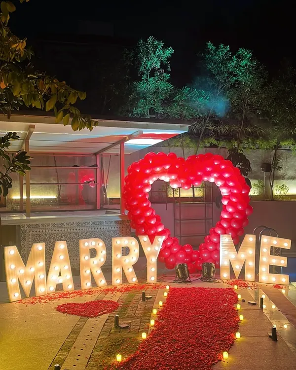 This poolside proposal setup features large, illuminated 'MARRY ME' letters and a dramatic heart-shaped balloon arch. A red petal-strewn walkway and ambient lighting create a truly cinematic and unforgettable moment.