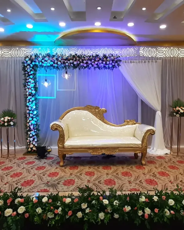 A sophisticated stage for an engagement ceremony. The decor features an L-shaped floral arrangement, elegant white drapery, and modern lighting fixtures, with a classic white and gold chaise lounge.