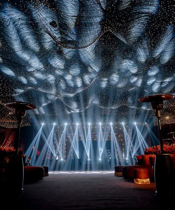 A breathtaking view of a sangeet stage set under a ceiling of thousands of fairy lights, creating a "starry night" effect. The powerful stage lighting and grand scale promise an evening of spectacular performances.