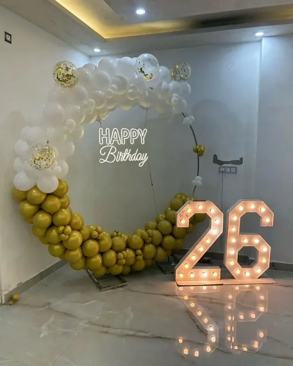 This gold and white theme is pure elegance. The setup features a half-and-half balloon ring, a 'Happy Birthday' neon sign, and large marquee numbers for the 26th birthday, all reflecting beautifully on the glossy floor.