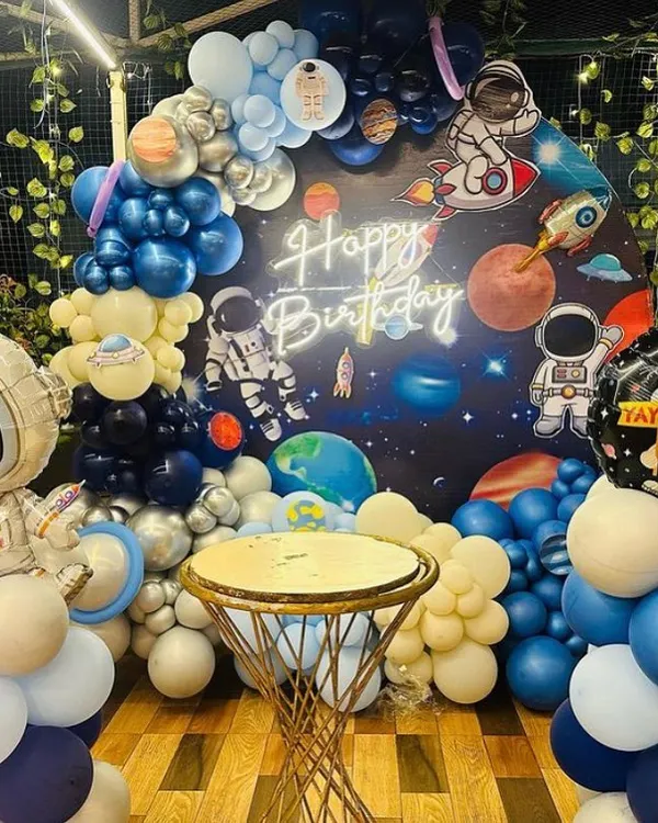 An out-of-this-world space theme party, perfect for a little astronaut's birthday. This setup features a galaxy backdrop with planets, a custom neon sign, and balloon garlands in shades of blue, silver, and white, complete with astronaut and rocket ship foil balloons.