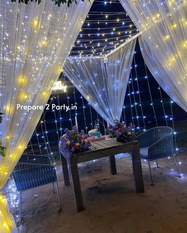 A classic romantic cabana dining setup for two, perfect for a surprise anniversary or birthday dinner. The structure is draped in sheer white curtains and wrapped in warm and cool fairy lights, creating a magical, star-like effect for your private meal.