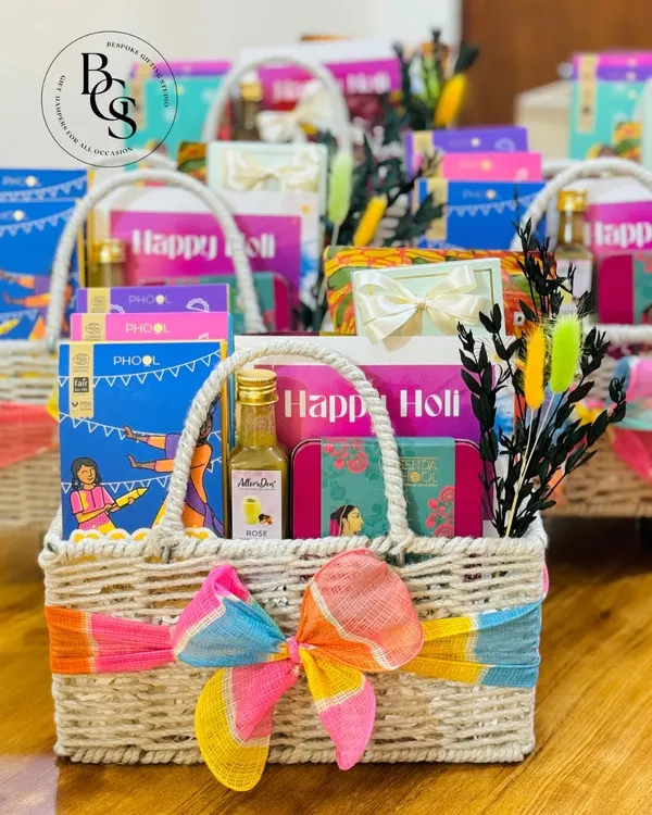 A vibrant Holi gift hamper in a woven basket. It's filled with festive essentials like organic colors from Phool, a bottle of rose sherbet, and other treats, all tied with a colorful tie-dye ribbon.