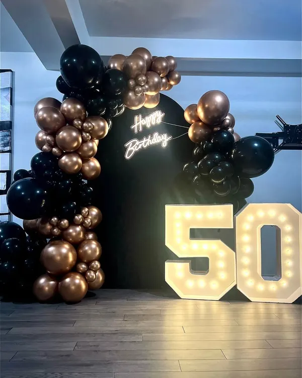 A sleek backdrop for a 50th birthday, featuring a black panel, a "Happy Birthday" neon sign, and large marquee numbers. The look is completed with a modern organic balloon arch in rose gold and black.