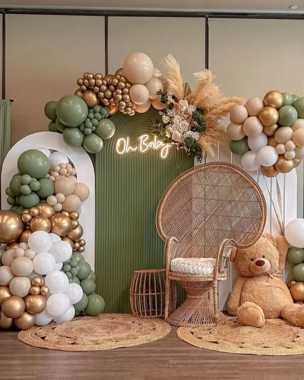 A stunning boho-chic baby shower with an olive green, white, and gold color scheme. This setup includes a wicker peacock chair, pampas grass, jute rugs, and an "Oh Baby" neon sign for a trendy and elegant look.