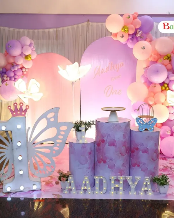 A beautiful butterfly-themed first birthday in shades of pink and purple. This setup includes two backdrops, organic balloon arrangements, light-up flower props, a large marquee number '1' with butterfly wings, and themed pedestals.