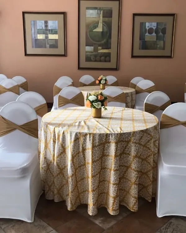 A clean and classic banquet seating arrangement featuring round tables with patterned cloths and elegant chair covers with gold sashes. This formal setup is ideal for corporate luncheons, seminars, and conferences.
