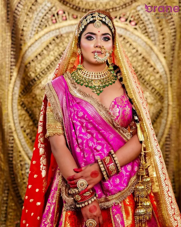 A regal bridal look featuring a multi-toned lehenga with pink and orange hues. The makeup is focused on bold eyes and is complemented by heavy, traditional jewelry.