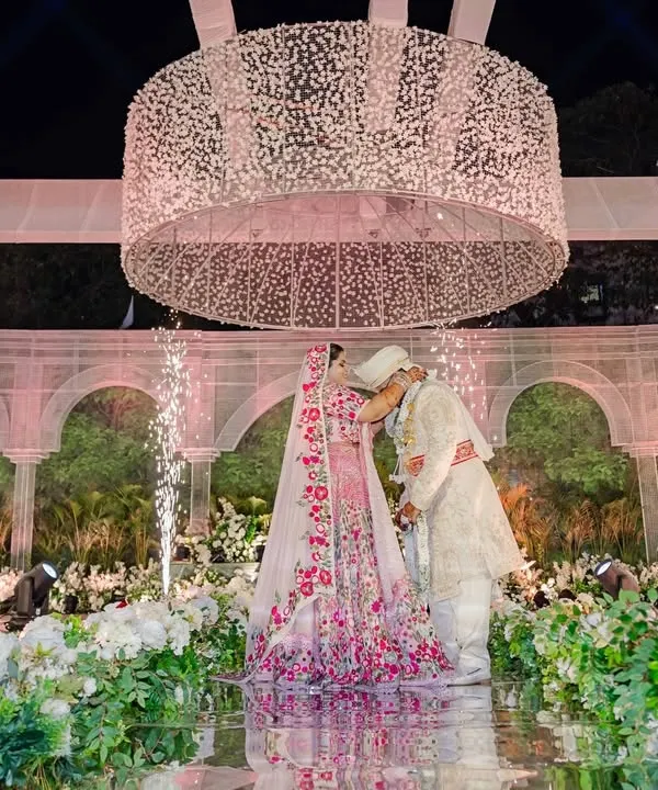 The pinnacle of romance: a couple shares a kiss under a massive, bespoke baby's breath chandelier. This breathtaking wedding moment is framed by a sustainable backdrop of living plants and elegant white arches, a testament to eco-luxe design.