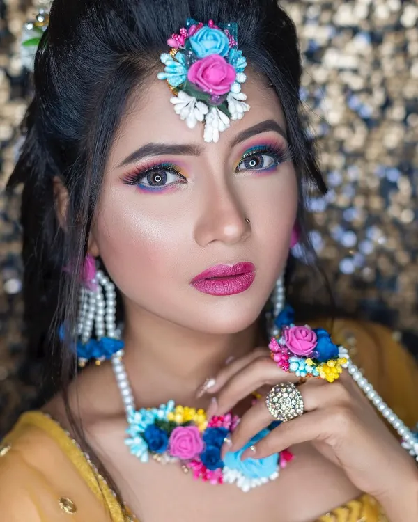 A colorful and creative look for a Haldi or Mehendi function. The eye makeup uses shades of blue and pink to match her floral jewelry, creating a unique and festive style.