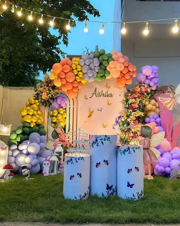 A stunning outdoor garden party with a butterfly theme. This elaborate setup features a massive, multi-colored balloon arch, floral arrangements, large butterfly props, and a light-up marquee number '5'.