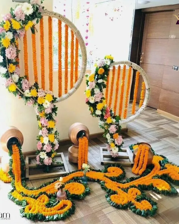 A creative and artistic floral installation for a home event. This decor features marigold garlands flowing from traditional pots, set against circular frames wrapped in flowers, creating a modern yet traditional look.
