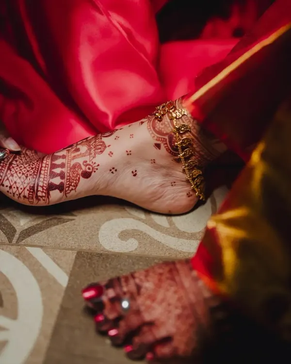 A candid shot of a beautiful henna stain on the foot, adorned with a golden anklet. The design features delicate birds and traditional patterns.