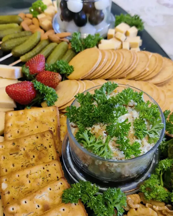 A beautiful cheese platter designed for a gathering. It features a variety of hard and soft cheeses, our homemade crackers, pickled gherkins, fresh strawberries, nuts, and a savory dip garnished with parsley.