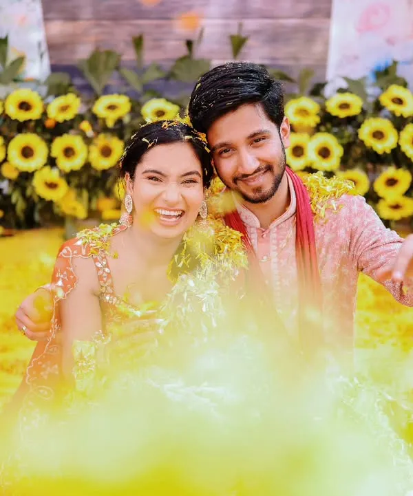 A fun and playful Haldi shot, with the couple smiling for the camera amidst a cloud of yellow turmeric powder.