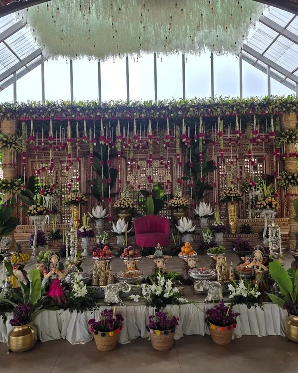 A complete view of a grand puberty ceremony stage I designed in a spacious venue with a glass roof. The decor features a massive floral backdrop with hanging tuberose and rose garlands, a central pink seat, and an elaborate display of traditional props and offerings.