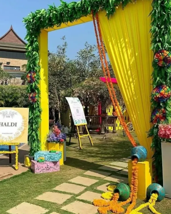 A welcoming entrance for a poolside Haldi event. This setup features a bright yellow arch with green foliage, a personalized "Haldi" welcome sign, and traditional pots with overflowing marigolds.