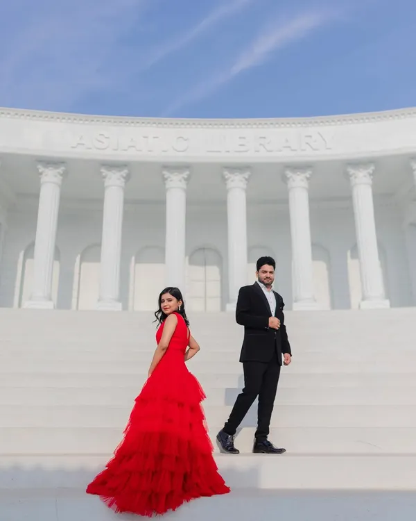 A dramatic, wide-angle shot on the steps of a grand, white library set. The bride's red gown creates a stunning visual pop, perfect for a movie-poster style pre-wedding photo.