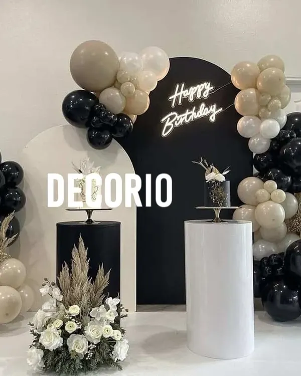 A modern and sophisticated birthday setup featuring a bold black circular backdrop with a custom 'Happy Birthday' neon sign. The scene is balanced with organic balloon garlands in black, white, and neutral tones, and includes elegant white and black pedestals for cakes.