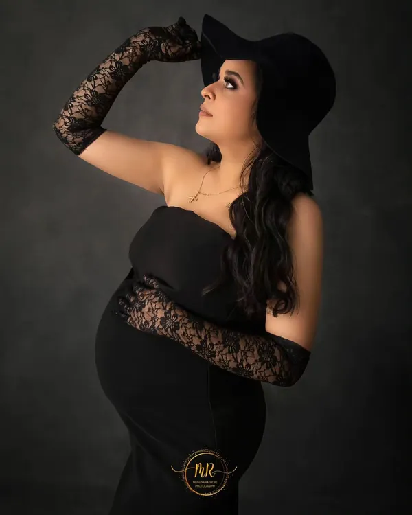 A chic and classic maternity portrait. The mother-to-be wears a simple black dress, elegant lace gloves, and a stylish hat for a timeless, fashionable look.