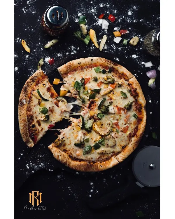 A top-down shot of a vegetable pizza, with a slice pulled away to show the cheese stretch. The dark, messy-on-purpose styling with scattered flour and toppings gives it an authentic, handcrafted feel.