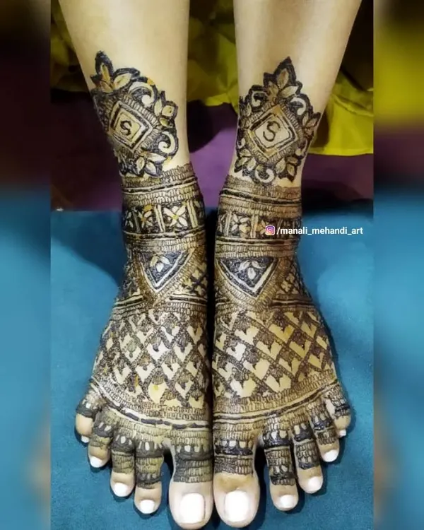 An intricate bridal mehandi design for the feet, featuring a dense net pattern and personalized initial 'S' motifs. The design covers the entire top of the foot and toes.
