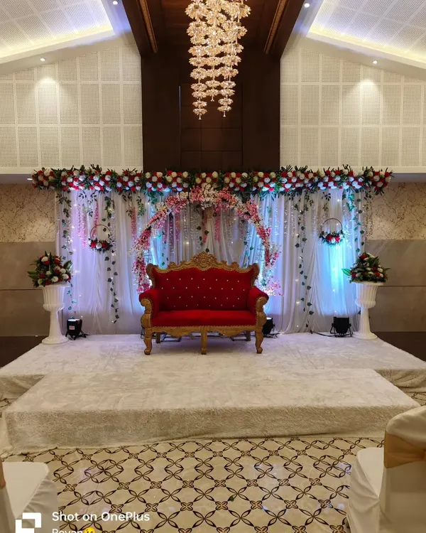 A beautiful reception stage setup in a modern banquet hall. The floral arch, soft drapery, and a classic red velvet sofa create a rich and romantic focal point for the couple.