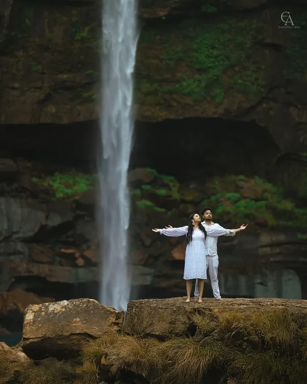 A couple stands on a rock ledge, arms outstretched, embracing the power of the waterfall behind them. This wide shot captures the scale of the landscape and the feeling of freedom.