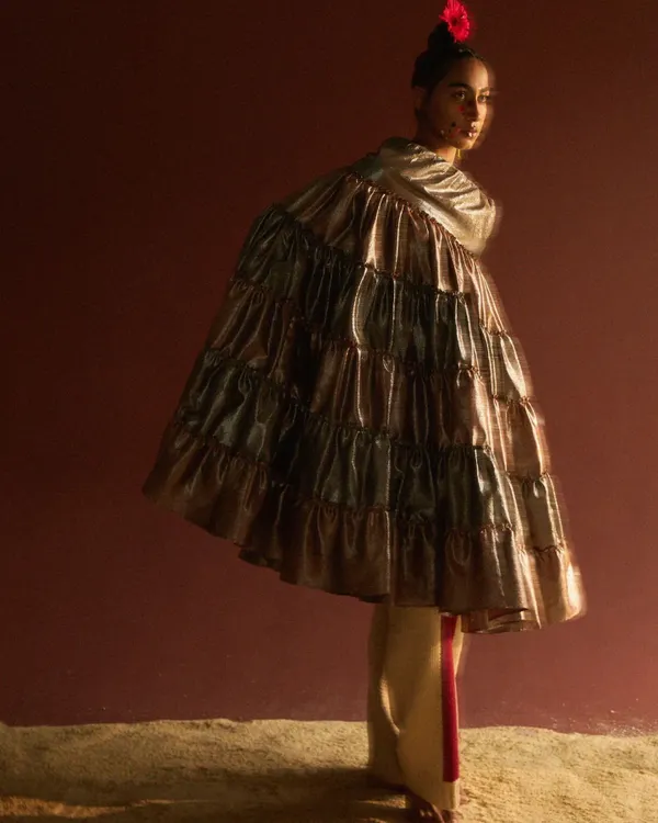 An avant-garde editorial look featuring a voluminous, tiered metallic cape. The styling creates a dramatic, sculptural silhouette, with a single red flower in the hair adding a point of sharp contrast against the moody, textured backdrop.